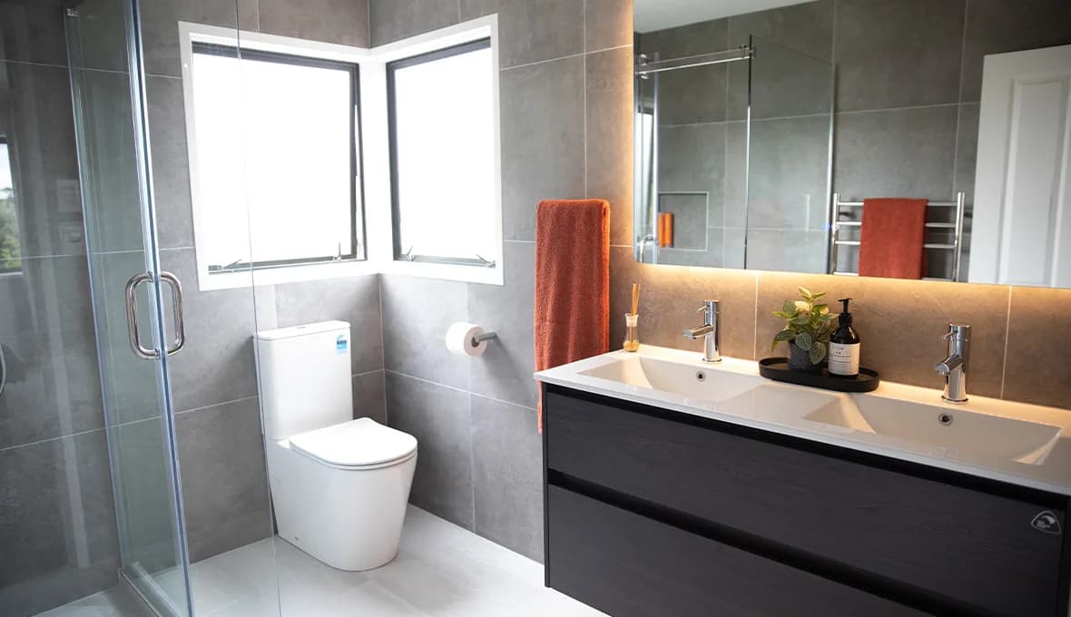 Grey stone-look bathroom with double vanity, two basins, matte black tapware, wall-hung toilet in a corner window nook, terracotta towel and frameless shower.