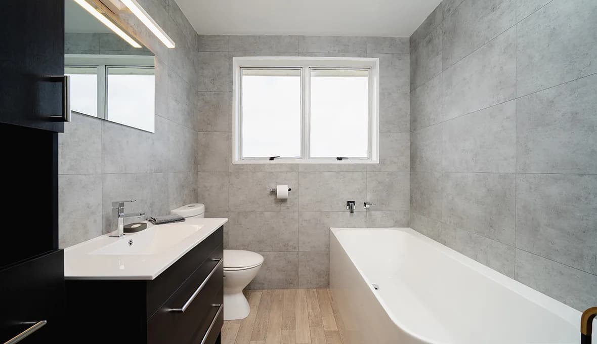 Bathroom with grey concrete-look wall tiles, timber-look floor tiles, dark timber wall-hung vanity, white top, chrome tap, wall-hung toilet and built-in bath.
