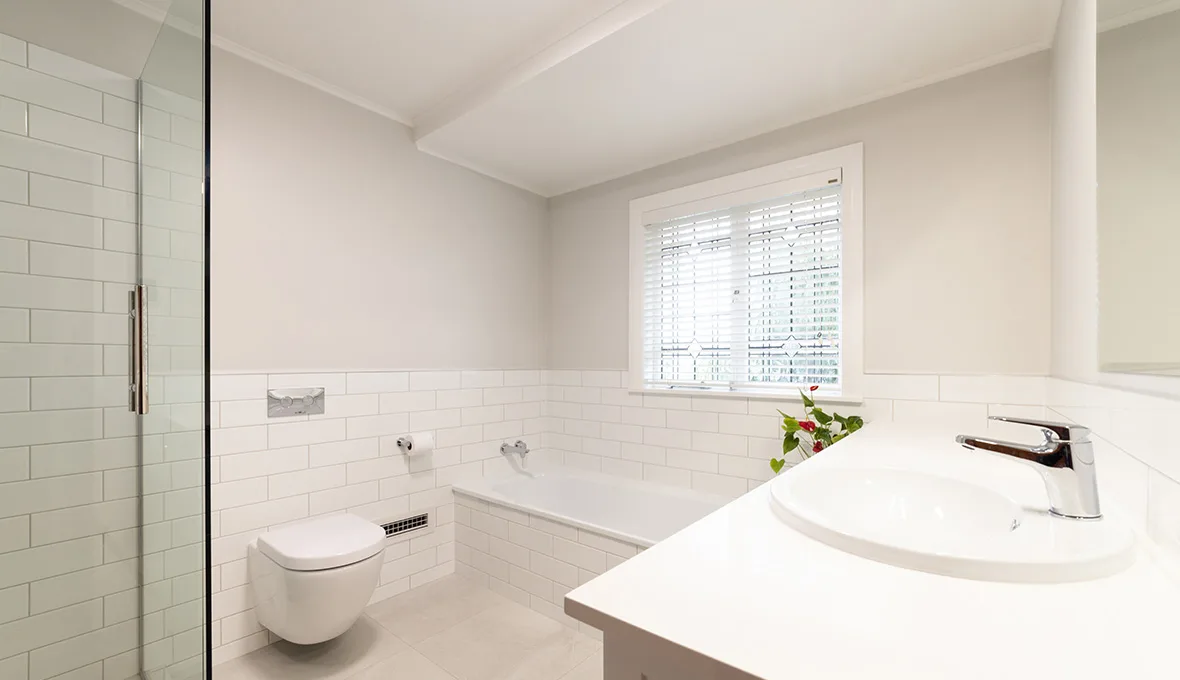 White subway-tile bathroom with frameless glass shower screen, wall-hung toilet, built-in bath below patterned blinds and white vanity with chrome tap.
