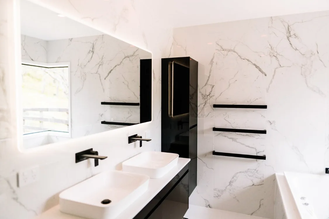 Bathroom with white marble-look large-format tiles and grey veining, a black gloss wall-hung vanity with a white stone top and two white rectangular vessel basins, matte black wall mixers, an LED backlit mirror, a tall black gloss storage tower, and black towel rails.