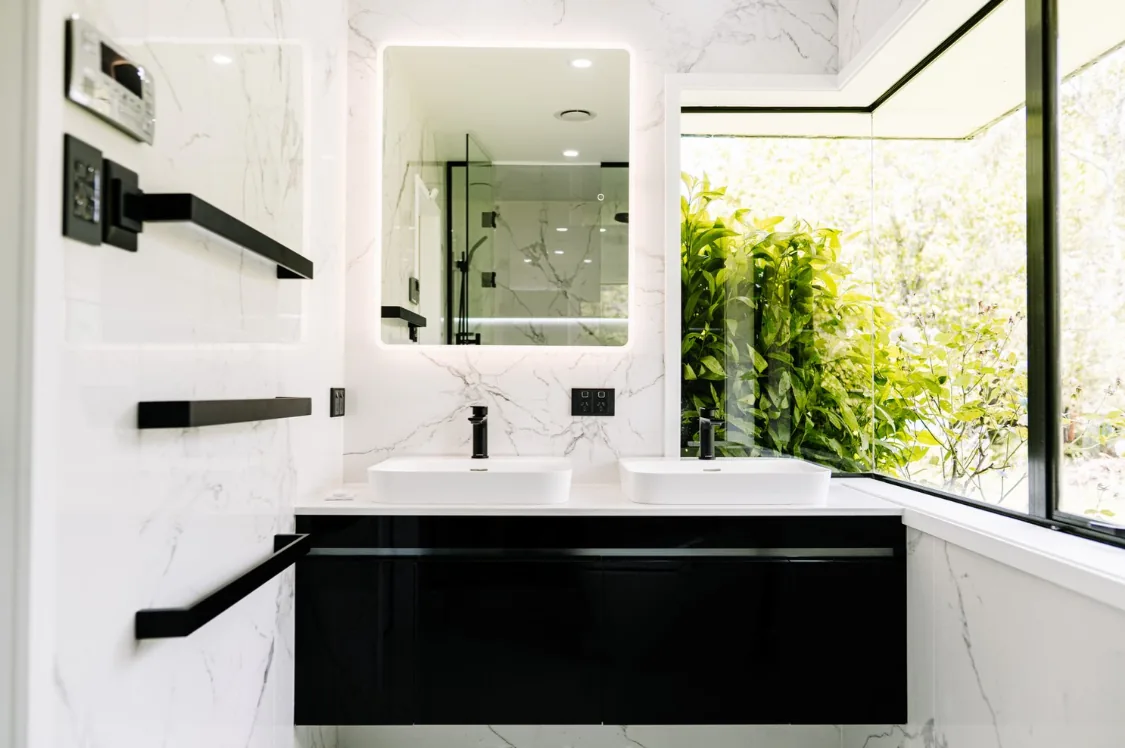 Ensuite with white marble-look tiles, black gloss wall-hung vanity, two white vessel basins, matte black tapware, LED mirror, black towel rails and corner window.