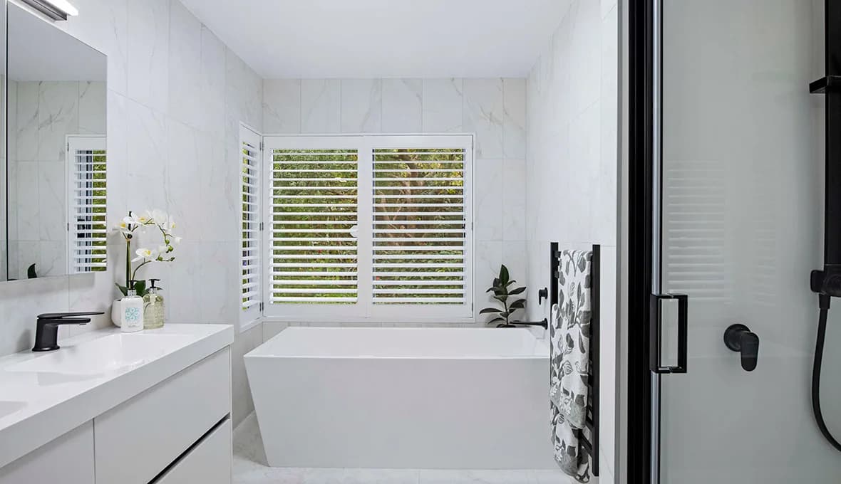 White freestanding bath beneath timber plantation shutters, with marble-look wall tiles, matte black ladder towel rail and frameless glass shower edge.