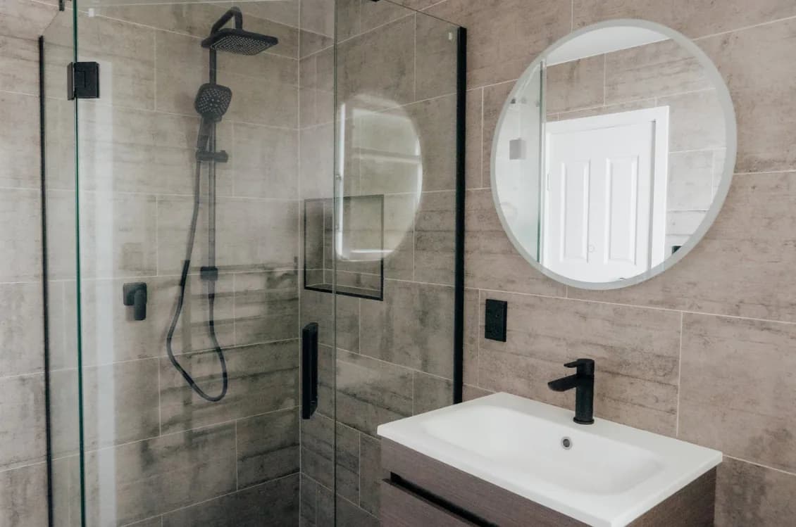 Warm grey bathroom with an angled black-framed corner shower, black rain head, timber-look wall-hung vanity, white basin and round LED mirror.