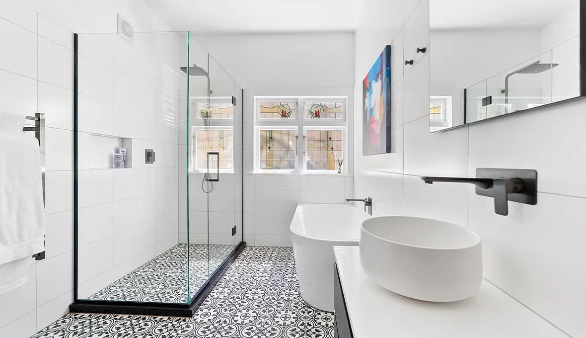 Bathroom with frameless glass shower, patterned floor tiles, original leadlight window, colourful portrait artwork and white freestanding bath with black spout.